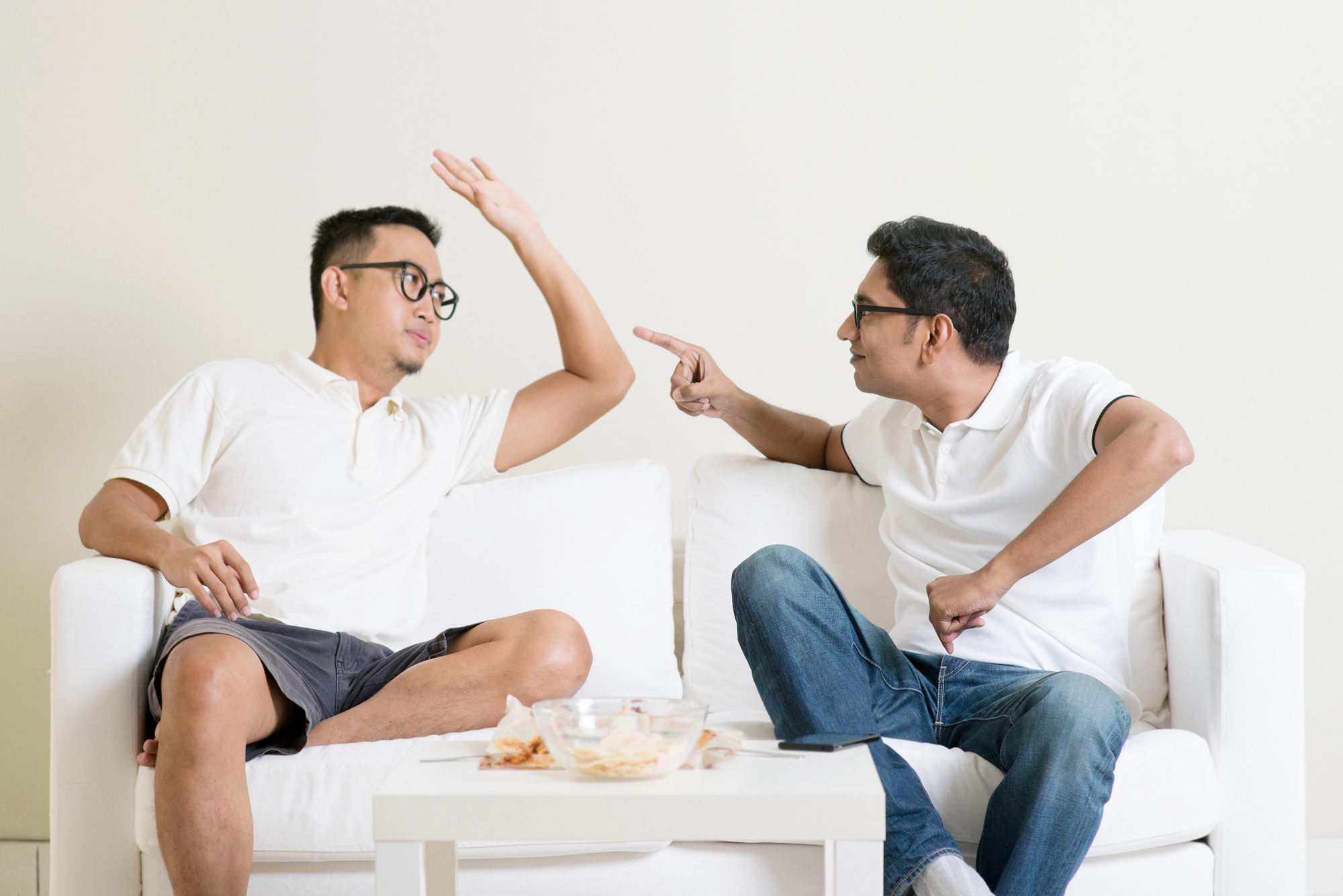 Two men sitting on a white couch engaging in a lively conversation. Both men wear glasses and casual white shirts; one is wearing shorts and the other jeans. The man on the left has his arm raised, while the man on the right points animatedly. A table with snacks sits in front of them.