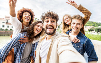 A group of five friends taking a selfie outdoors. They are all smiling and appear joyful. One person is giving a piggyback ride, while another is making a peace sign with their hand. They are standing against a backdrop of buildings and greenery.