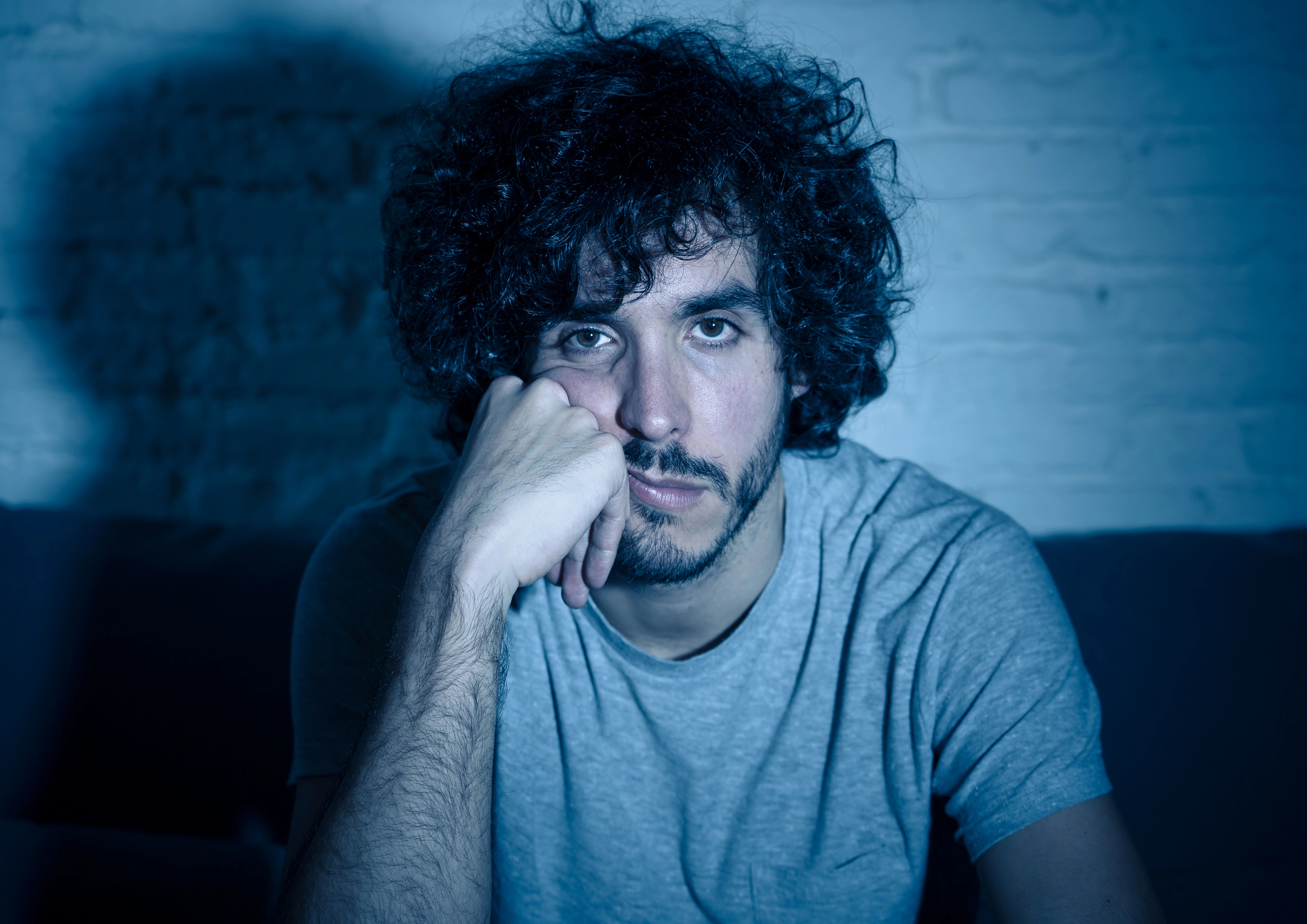 A man with curly hair and a beard is sitting with his chin resting on his left hand. He is wearing a gray t-shirt and has a serious, contemplative expression. The background is a brick wall, and the image is cast in blue-toned lighting.