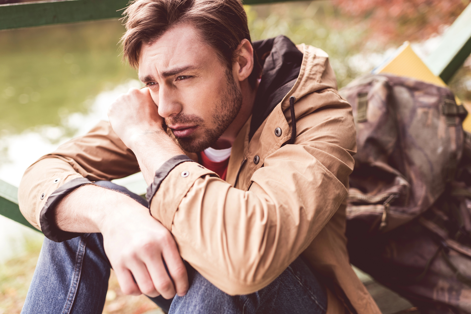 A young man with short brown hair, wearing a beige jacket and blue jeans, sits outdoors by a body of water, looking thoughtful and resting his head on his arm. A camouflage-patterned backpack is beside him. Fall foliage is visible in the background.
.