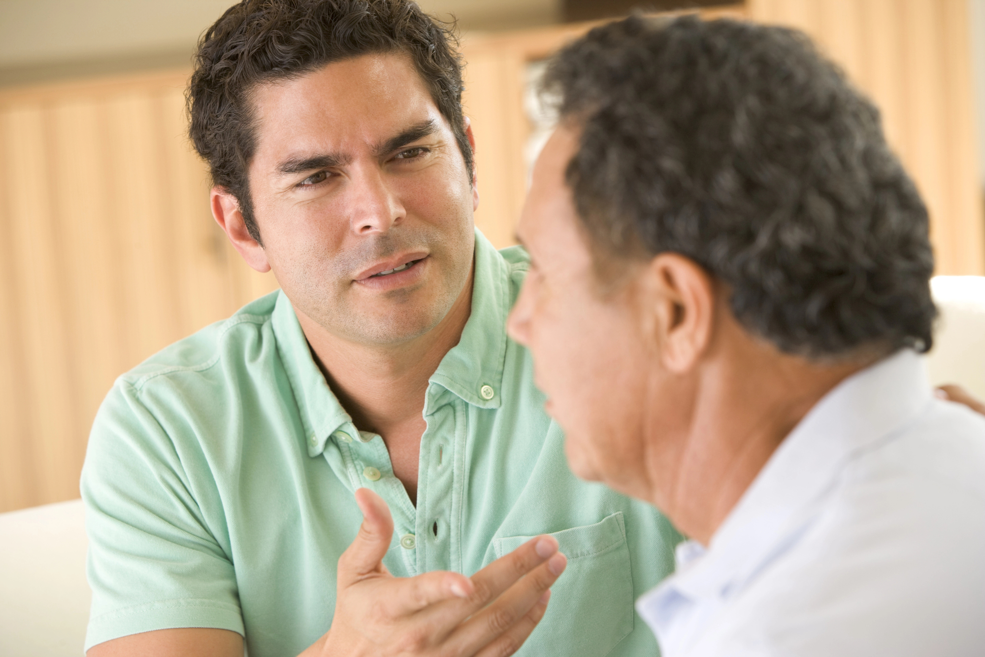 A man with short, dark hair, wearing a light green shirt, is engaged in a serious conversation with an older man who has gray hair and is wearing a light blue shirt. The younger man is gesturing with his hand as he speaks.