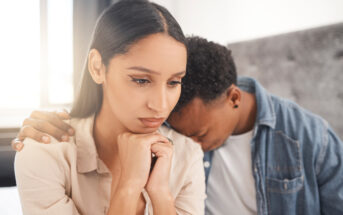 A woman with a thoughtful expression rests her chin on her hands, while a man sitting next to her leans his head on her shoulder and places his hand on her upper arm. They appear to be in a comforting and supportive moment.