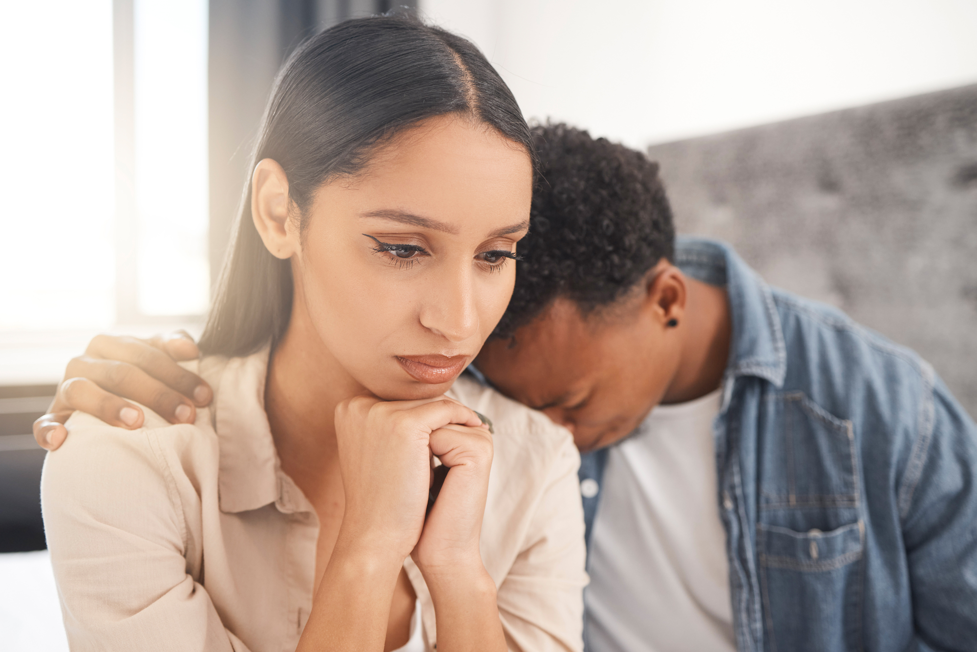 A woman with a thoughtful expression rests her chin on her hands, while a man sitting next to her leans his head on her shoulder and places his hand on her upper arm. They appear to be in a comforting and supportive moment.