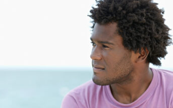 A person with short curly hair and a beard is gazing thoughtfully to the side. They are wearing a pink shirt and the background shows a blurred view of the ocean under a clear sky.