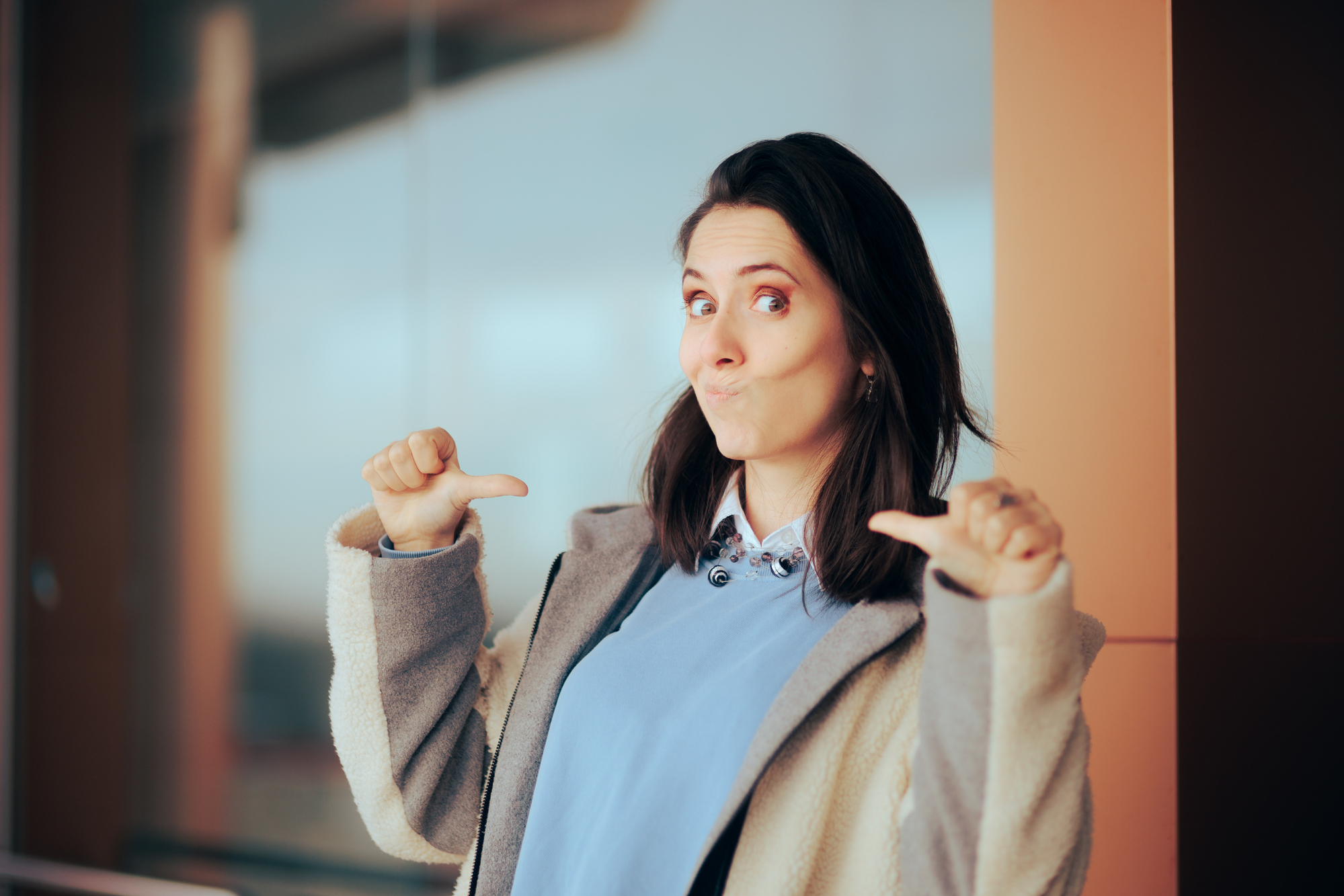 A woman with shoulder-length dark hair smiles and points both thumbs at herself in a playful manner. She is wearing a light gray coat over a blue top and stands in front of a blurred background featuring glass and neutral-colored structures.