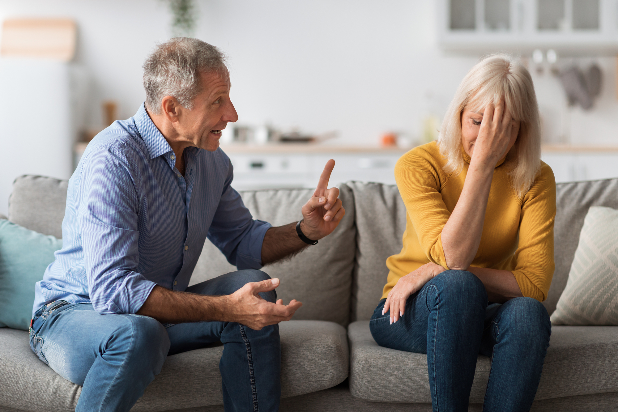 An elderly man and woman sit on a couch in a modern living room. The man, wearing a blue shirt, is speaking animatedly with one finger raised. The woman, wearing a yellow sweater, rests her head in her hand, appearing frustrated or distressed.