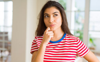 A woman with long dark hair wearing a red and white striped shirt is standing indoors, thoughtfully placing her hand on her chin. She looks upwards as if pondering something. The background is blurred, showing a well-lit room with windows.