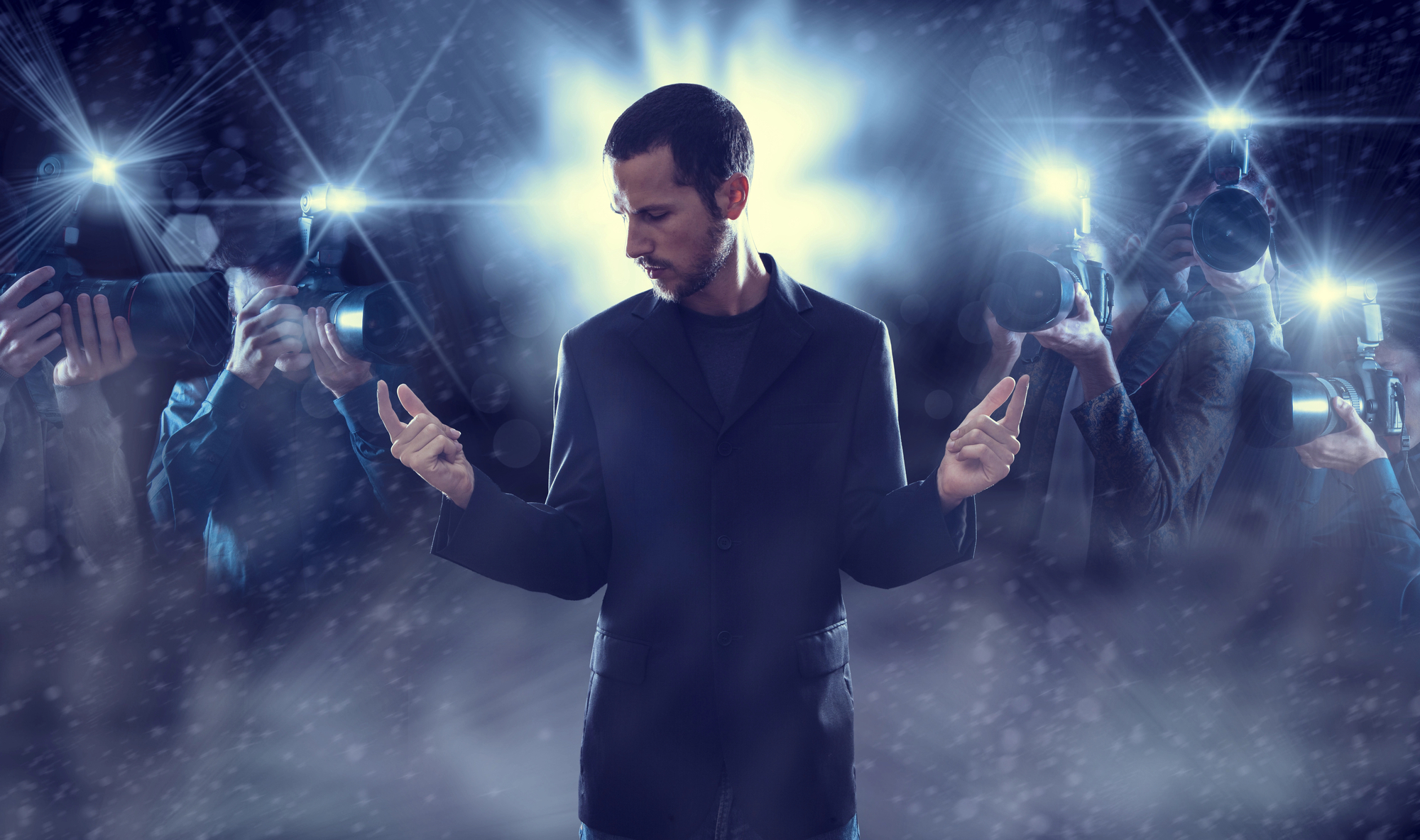 A man stands in a spotlight wearing a black suit jacket, looking down and pointing his fingers outward. Bright lights and camera flashes surround him, capturing his image from various angles, creating a sense of fame and attention. The background is dark and blurred.