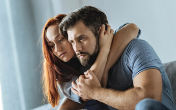 A woman with long red hair embraces a bearded man wearing a blue striped shirt, both looking contemplative and serious. They are indoors, in a well-lit room with a calm, soft background. The scene conveys a sense of comfort and support.