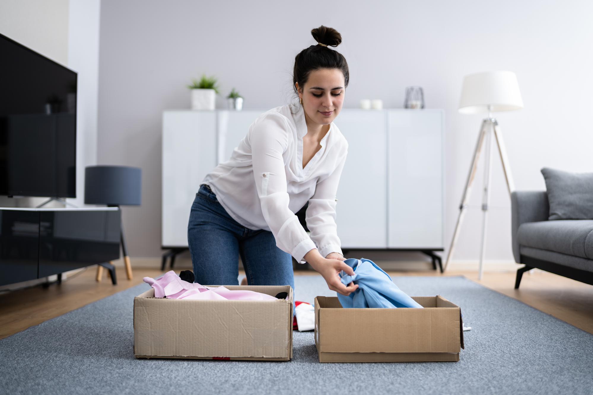 A woman with dark hair tied in a bun is kneeling on a grey carpet in a living room, organizing clothes from cardboard boxes. She is wearing a white blouse and blue jeans. The room has modern furniture, including a lamp, a TV, and a grey sofa.