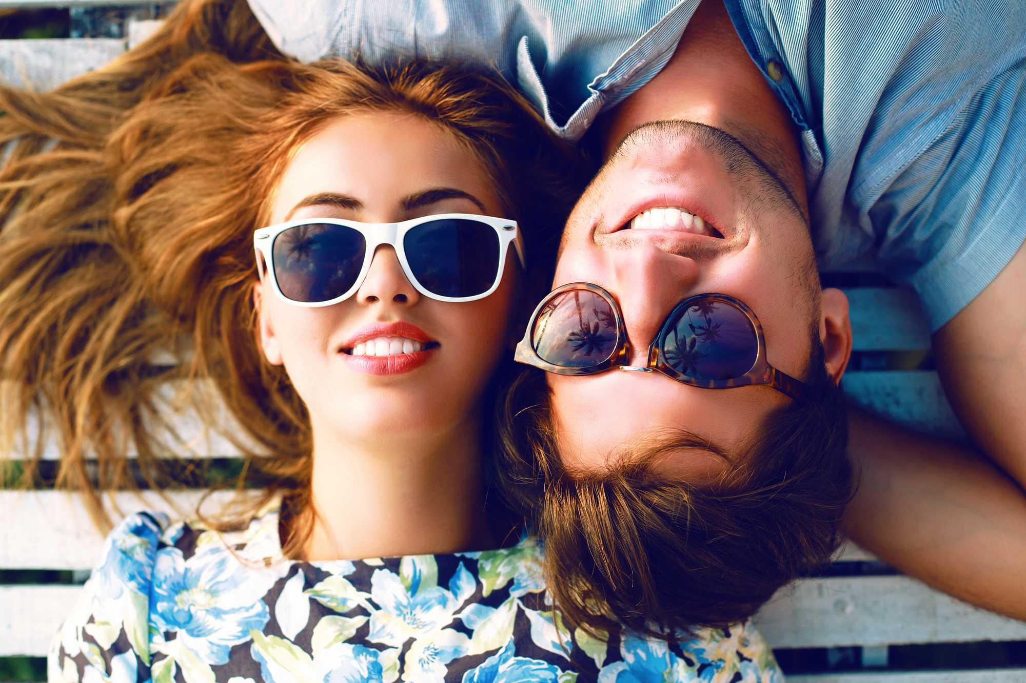 A young woman wearing white sunglasses and a floral dress lies beside a young man wearing dark sunglasses and a light blue shirt. They both smile, lying on a wooden surface with heads together but in opposite directions. The photo is taken from above.