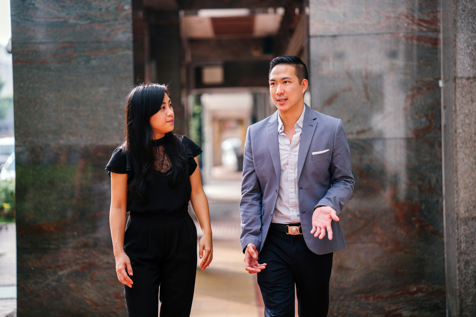 A man and woman are walking and having a conversation. The man, dressed in a light-colored shirt, dark pants, and a gray blazer, gestures with one hand while speaking. The woman, dressed in black, listens attentively. They are in an outdoor urban setting with stone walls.