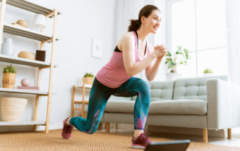 A woman in exercise clothes does lunges in a bright living room with a laptop in front of her. She is wearing a pink tank top and colorful leggings. The room has a sofa, wire shelf with decorative items, and a large window letting in natural light.