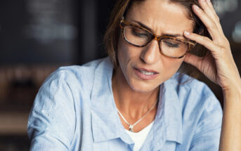 A woman with glasses and brown hair, wearing a light blue shirt and a necklace, holds her head with one hand and looks down with a worried expression. The background is blurred.