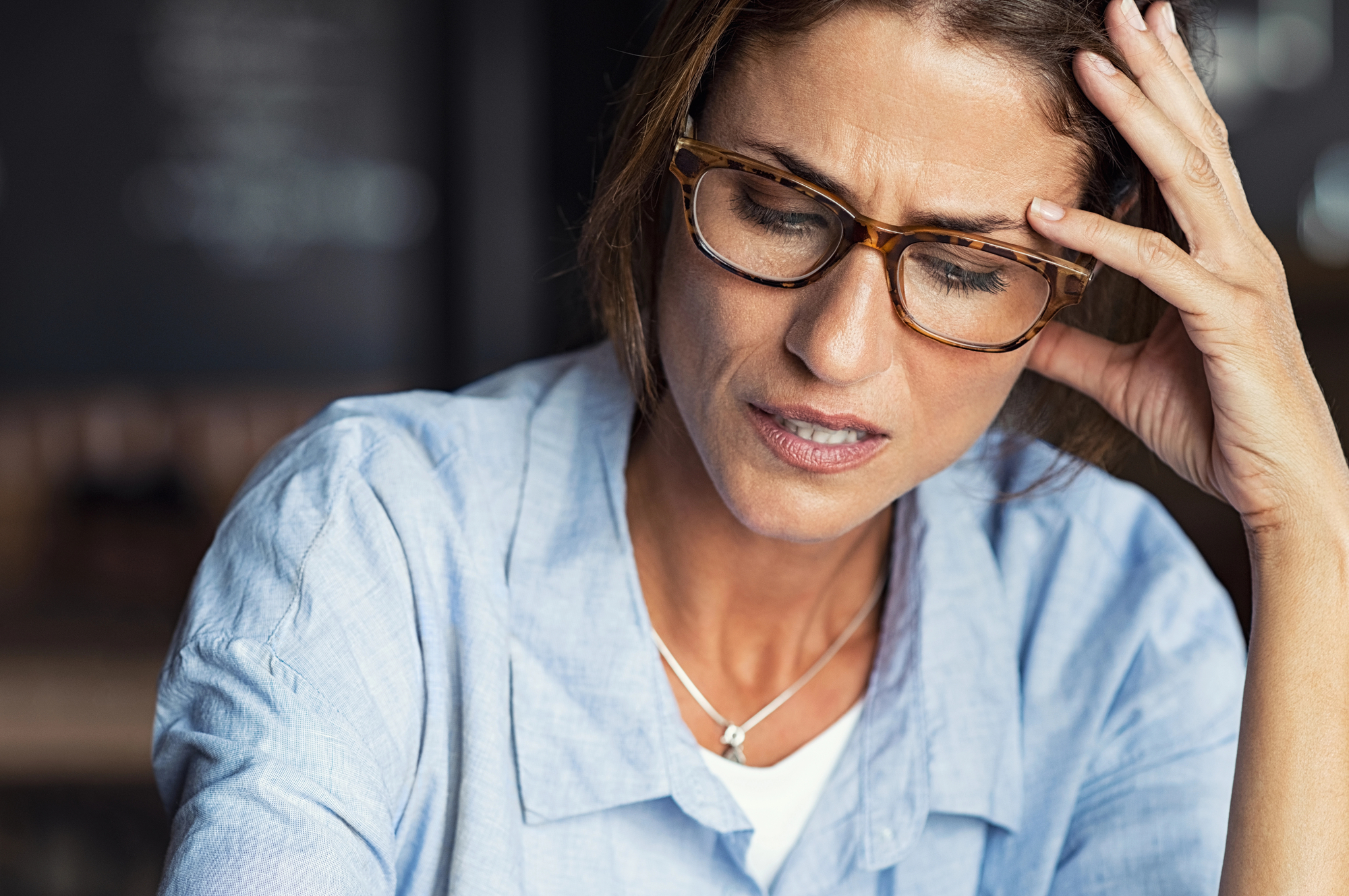 A woman with glasses and brown hair, wearing a light blue shirt and a necklace, holds her head with one hand and looks down with a worried expression. The background is blurred.