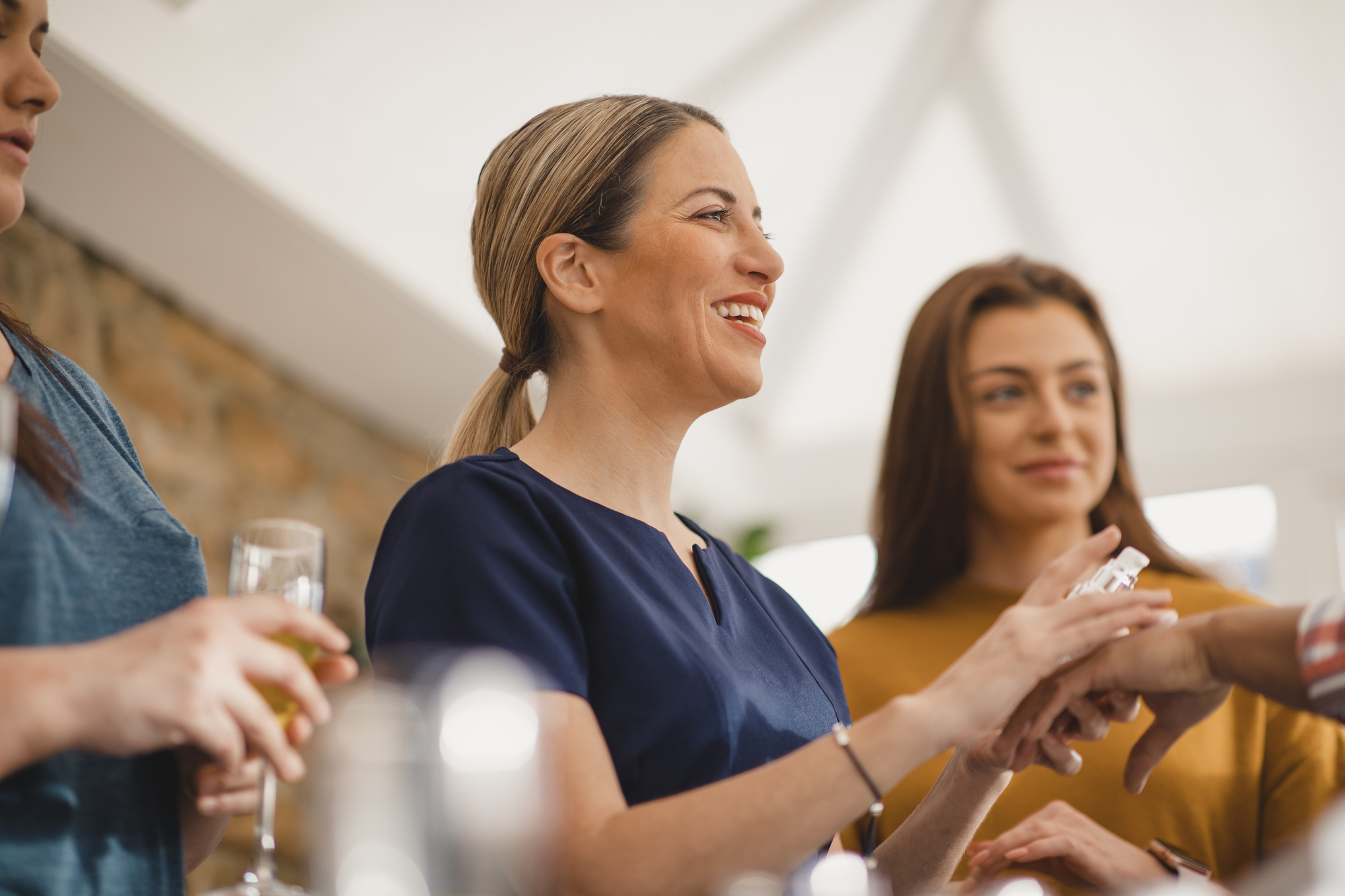 A smiling woman with her hair tied back in a ponytail is gesturing with her hands. She is surrounded by others who are partially visible, one holding a glass. The group appears to be indoors, engaged in a friendly conversation or gathering.