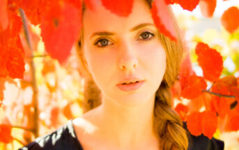 A woman with long hair in a braid stands amidst bright red autumn leaves. She gazes directly at the camera with a calm expression, her face softly lit by the sunlight filtering through the foliage. She wears a black top, and the vibrant leaves frame her face.