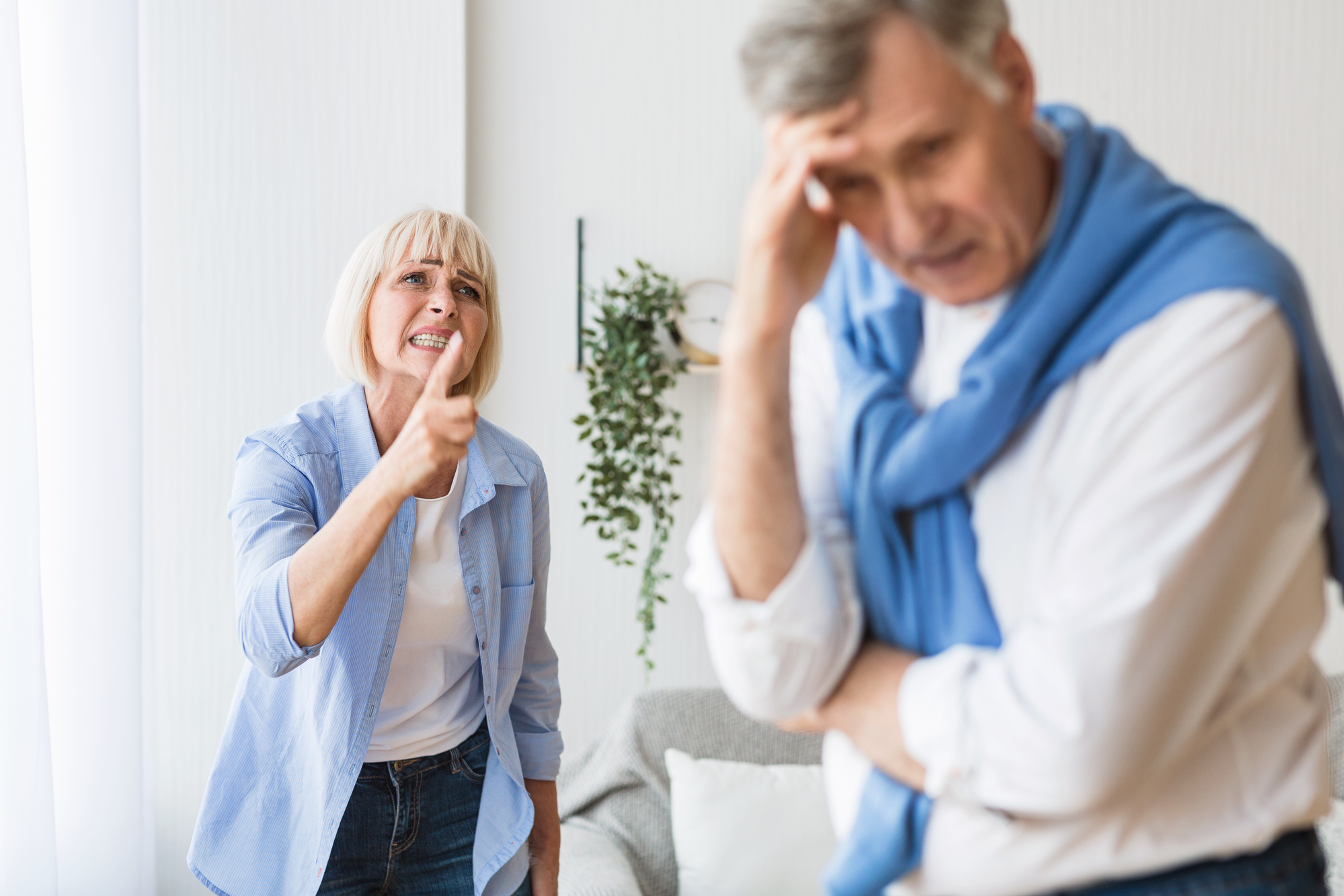 An older woman with short blonde hair is angrily pointing a finger and yelling at an older man. The man, with graying hair, appears distressed, holding his hand to his forehead. They are in a light-colored room with a plant hanging in the background.