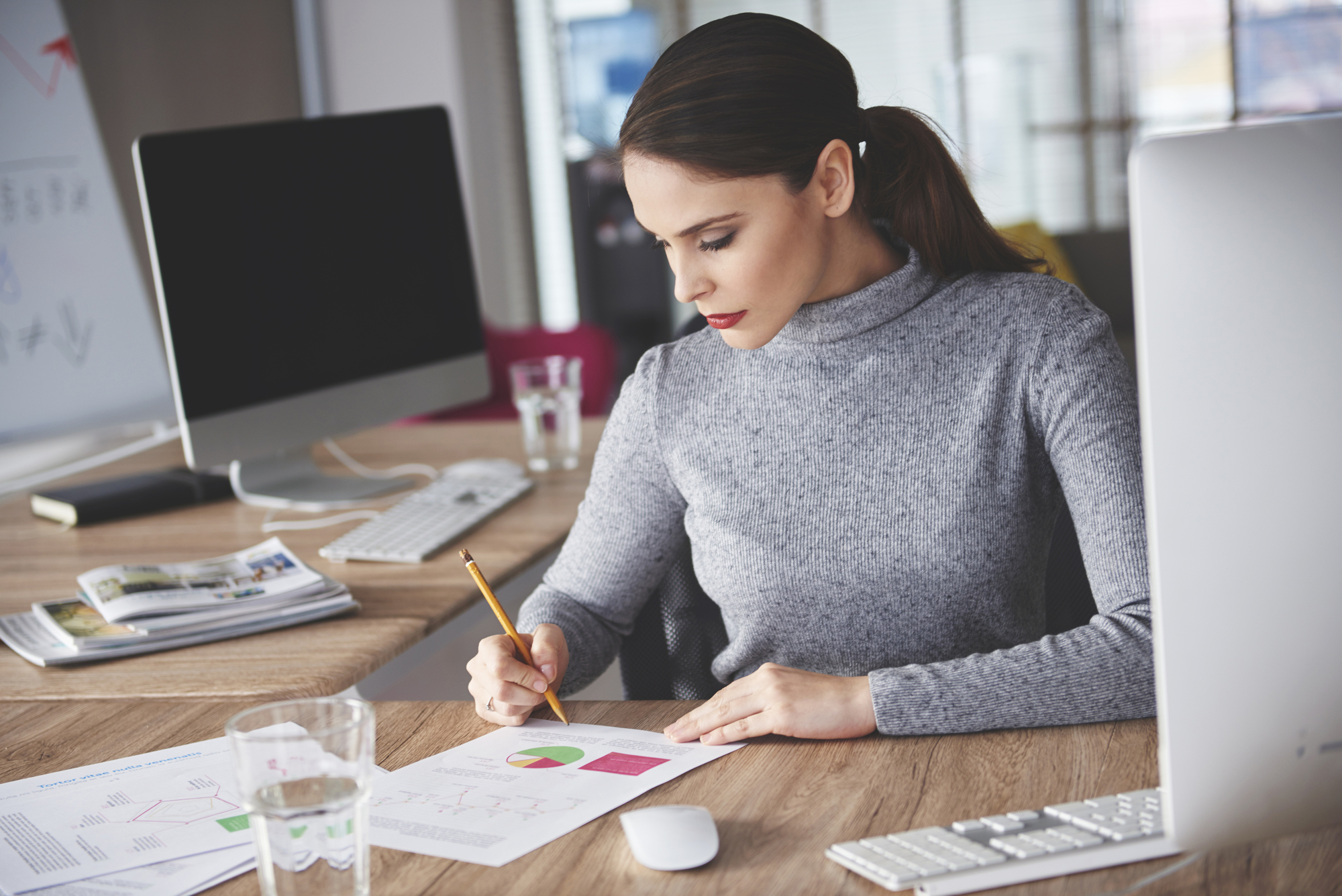 A woman with dark hair, tied in a ponytail, is seated at a wooden desk in an office, analyzing a chart with a pencil. Two computer monitors, a keyboard, a mouse, a glass of water, and open documents are on the desk. The background shows bookshelves and large windows.