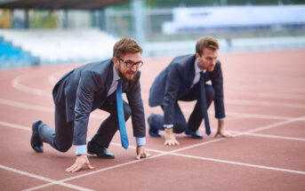 Two men in business suits and ties crouching in starting positions on a running track, ready to race. The background shows a blurred stadium with empty seating. The image symbolizes competition in the business world.