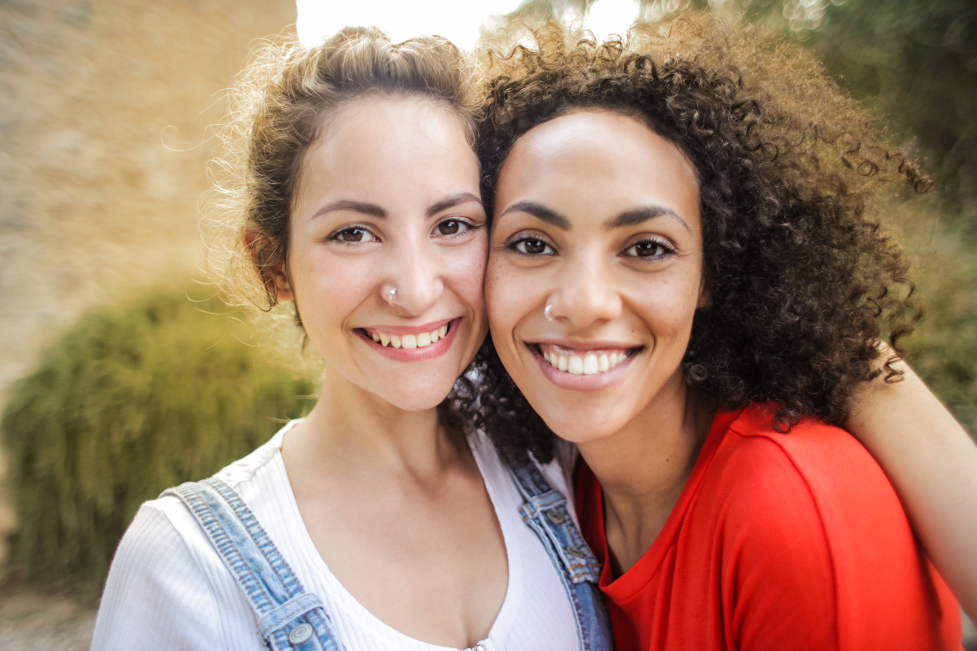 Two women are smiling and posing closely for a photo outdoors. One woman is wearing a white top with denim overalls, and the other is in a red top. They both have curly hair and look happy. The background is blurred with greenery and a building visible.