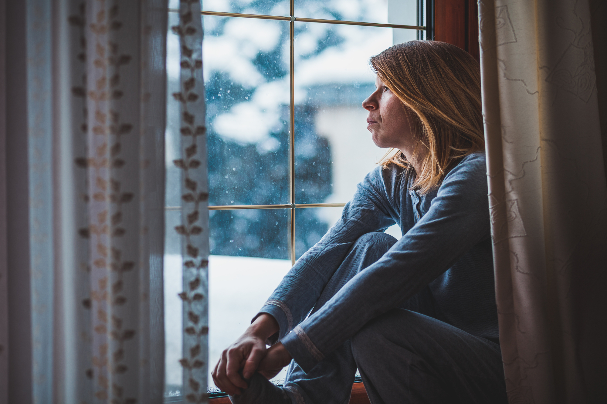 A woman in blue pajamas sits by a window, looking out at a snowy landscape. She appears deep in thought, with her hands resting on her knees and her head slightly tilted to the side. The cold winter scene outside contrasts with the cozy interior.