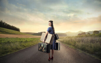 A woman in a blue polka dot dress and red shoes stands on a rural road at sunset, holding two vintage suitcases and looking back over her shoulder. Green fields and rolling hills stretch out on either side under a partly cloudy sky.