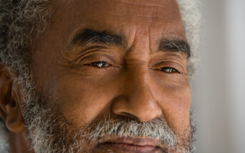 Close-up portrait of an elderly man with gray hair and a beard. He has a calm expression with slightly closed eyes, showcasing the fine wrinkles on his face. The background is blurred, keeping the focus on his contemplative gaze and facial details.