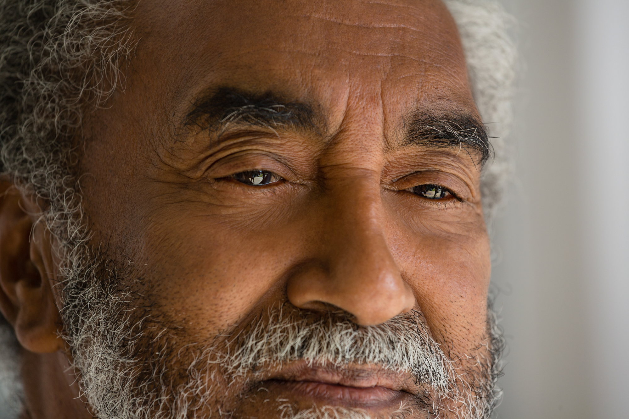 Close-up portrait of an elderly man with gray hair and a beard. He has a calm expression with slightly closed eyes, showcasing the fine wrinkles on his face. The background is blurred, keeping the focus on his contemplative gaze and facial details.