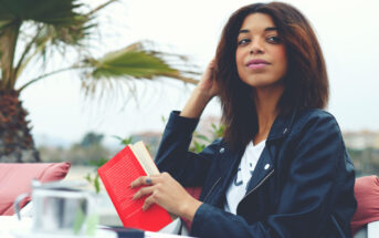 A woman with shoulder-length dark hair, wearing a black leather jacket, is seated outdoors at a table with a red book in her hand. She looks slightly to her right, with palm trees in the background, and a teapot and cup on the table in front of her.