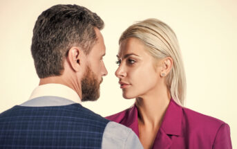 A man and a woman stand facing each other closely, gazing into each other’s eyes. The man has short, dark hair with a beard and wears a checkered suit jacket over a white shirt. The woman has long, blonde hair pulled back and wears a magenta blazer.