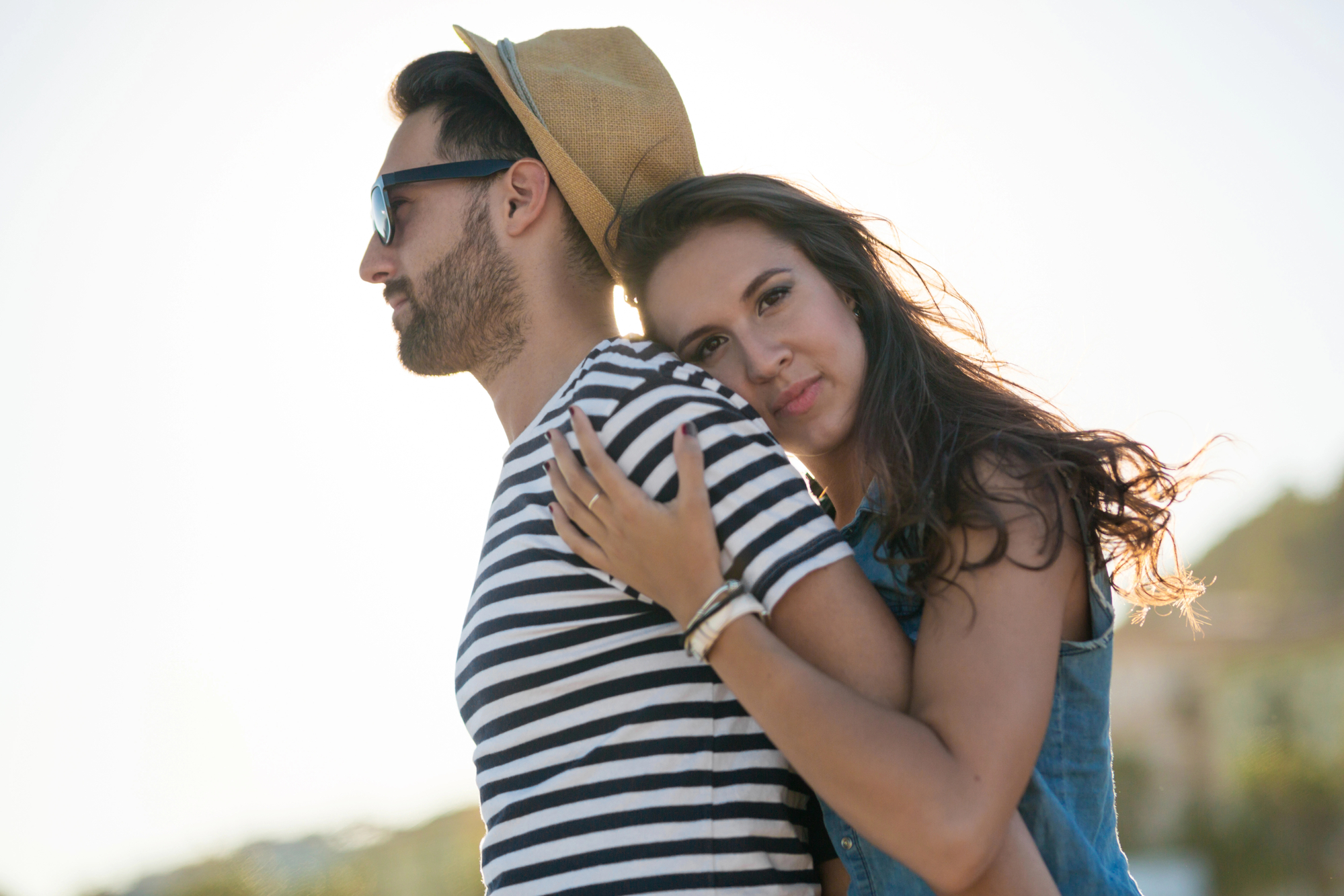 A young woman embraces a young man from behind. The man wears a striped shirt, sunglasses, and a hat. The woman is wearing a denim top and has her arms around his shoulders. They are outdoors, with a blurred background of trees and a bright sky.