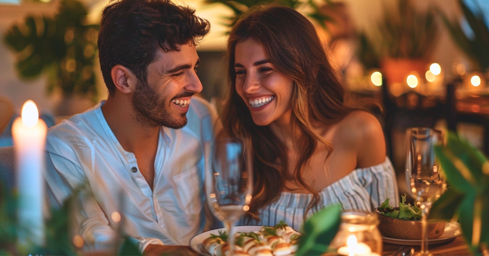 A couple sits at a table enjoying a romantic dinner with soft candlelight. They are smiling at each other, surrounded by plants and elegant table settings, creating a warm and intimate atmosphere.