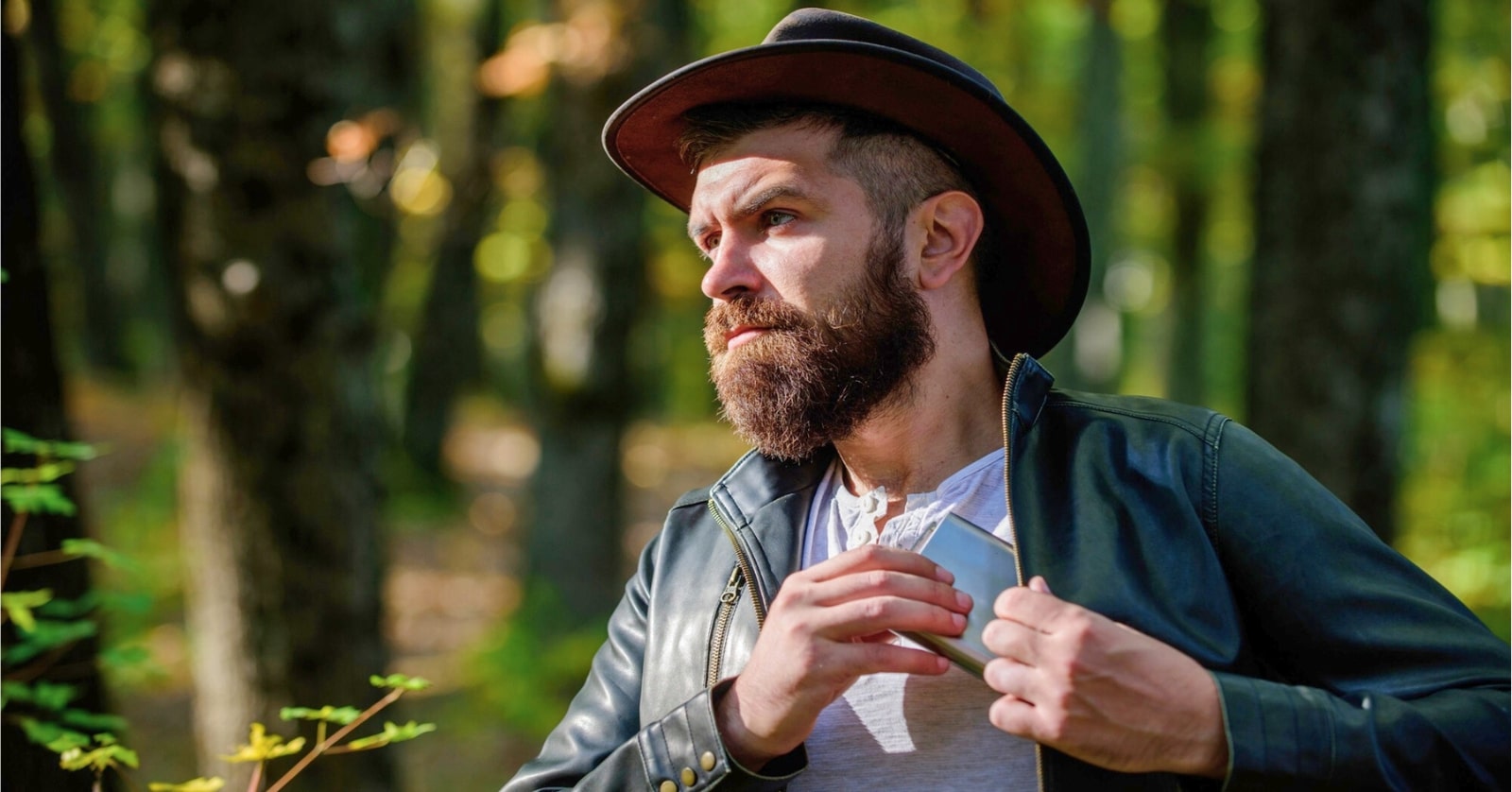 A bearded man wearing a wide-brimmed hat and a leather jacket stands in a forest while looking to his left. He is holding a folded map and appears to be preparing to unfold it. Sunlight filters through the trees, casting dappled light on the scene.
