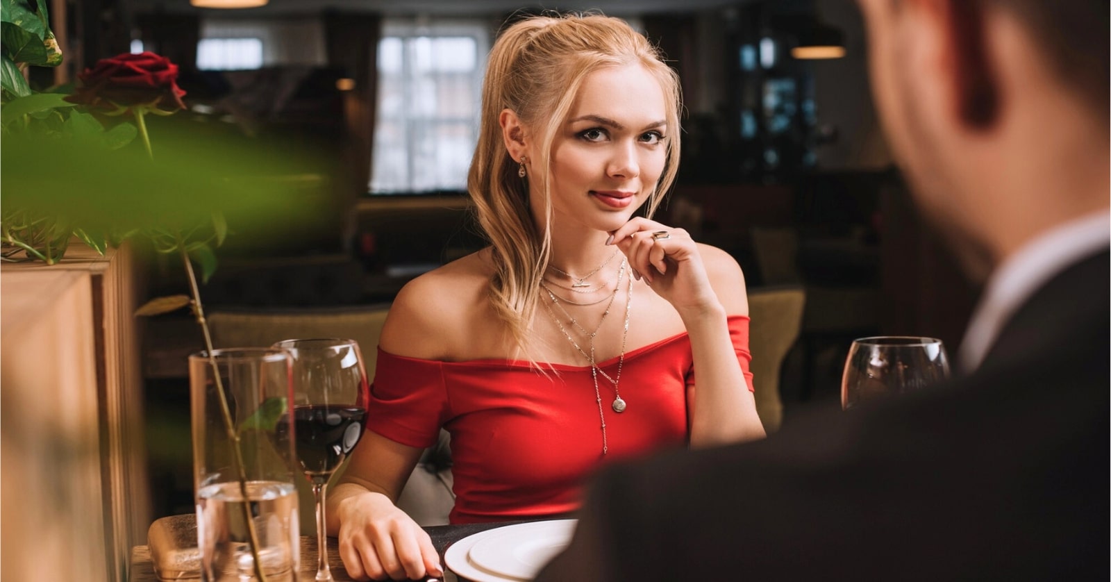 A woman with blonde hair, tied in a ponytail, sits at a restaurant table adorned with wine glasses, a red rose, and plates. She wears a red off-shoulder top and jewelry, and looks across the table with a soft smile, while a man, slightly blurred, faces her.