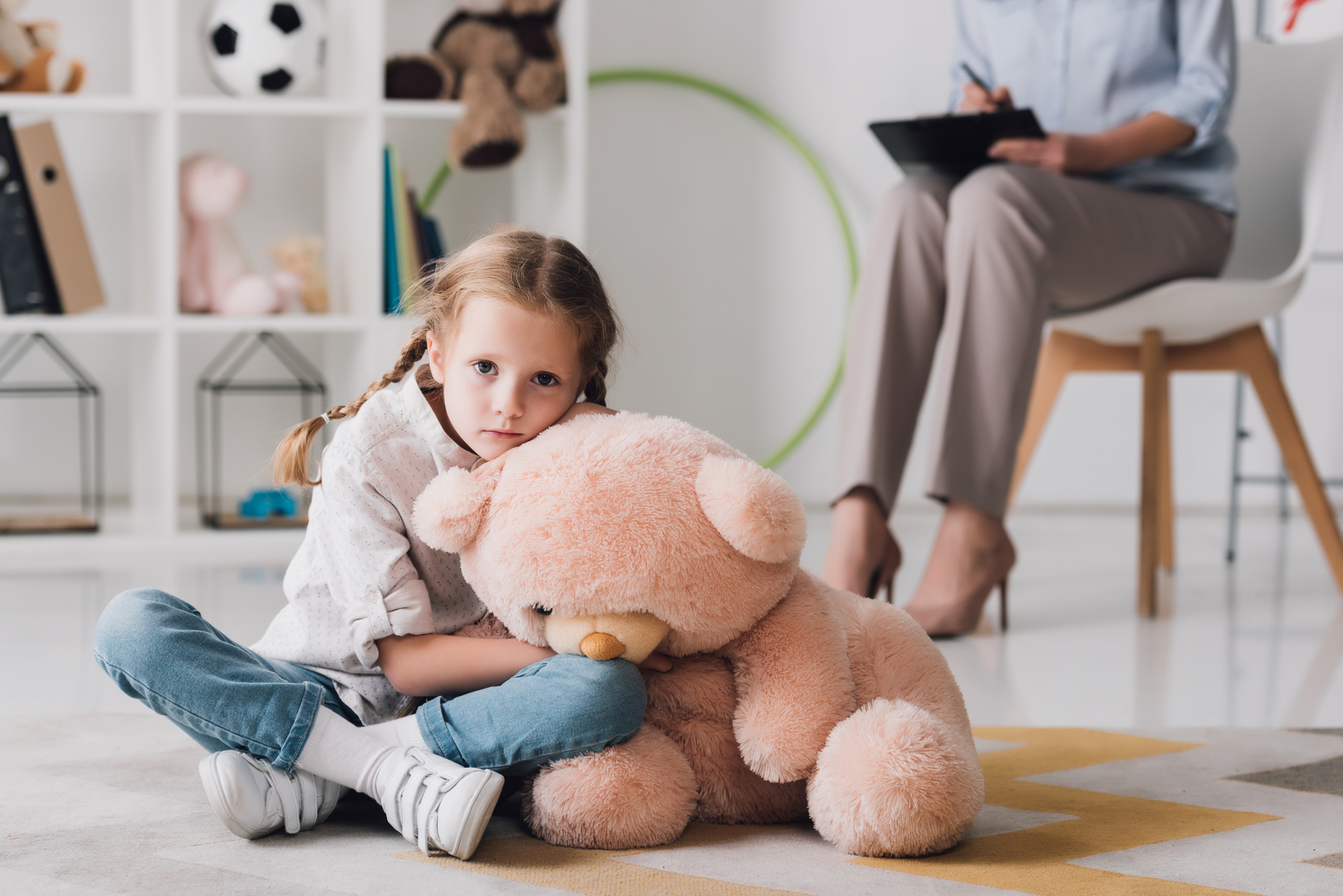 A young girl with pigtails sits on the floor hugging a large stuffed teddy bear, looking sad. In the background, an adult sits on a chair with a clipboard, suggesting a therapy or counseling session. Shelves with toys and books are visible in the room.