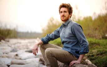 A man with curly hair and a beard is sitting on a large rock beside a riverbank. He is wearing a blue buttoned-down shirt with rolled-up sleeves and khaki pants. The background features trees and a cloudy sky. The scene exudes a calm, contemplative mood.