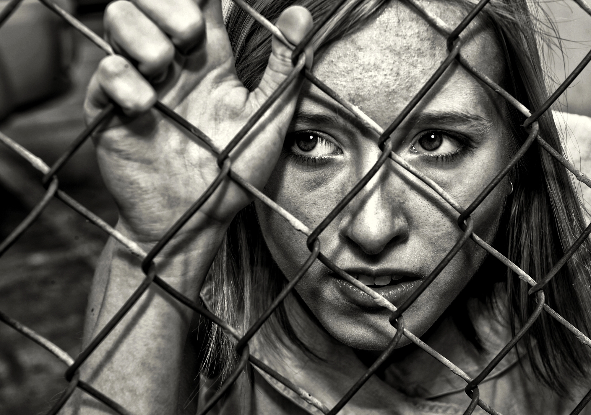 A close-up black and white photo of a girl looking through a chain-link fence. She has a worried expression, gripping the fence with one hand. Her face shows dirt and smudges, reflecting a tough or challenging situation.