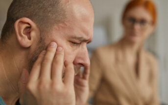 A man with a bald head and facial hair sits with his eyes closed and fingers on his temples, looking stressed or in pain. In the blurred background, a woman with red hair and glasses observes him attentively. Their expressions suggest concern and empathy.