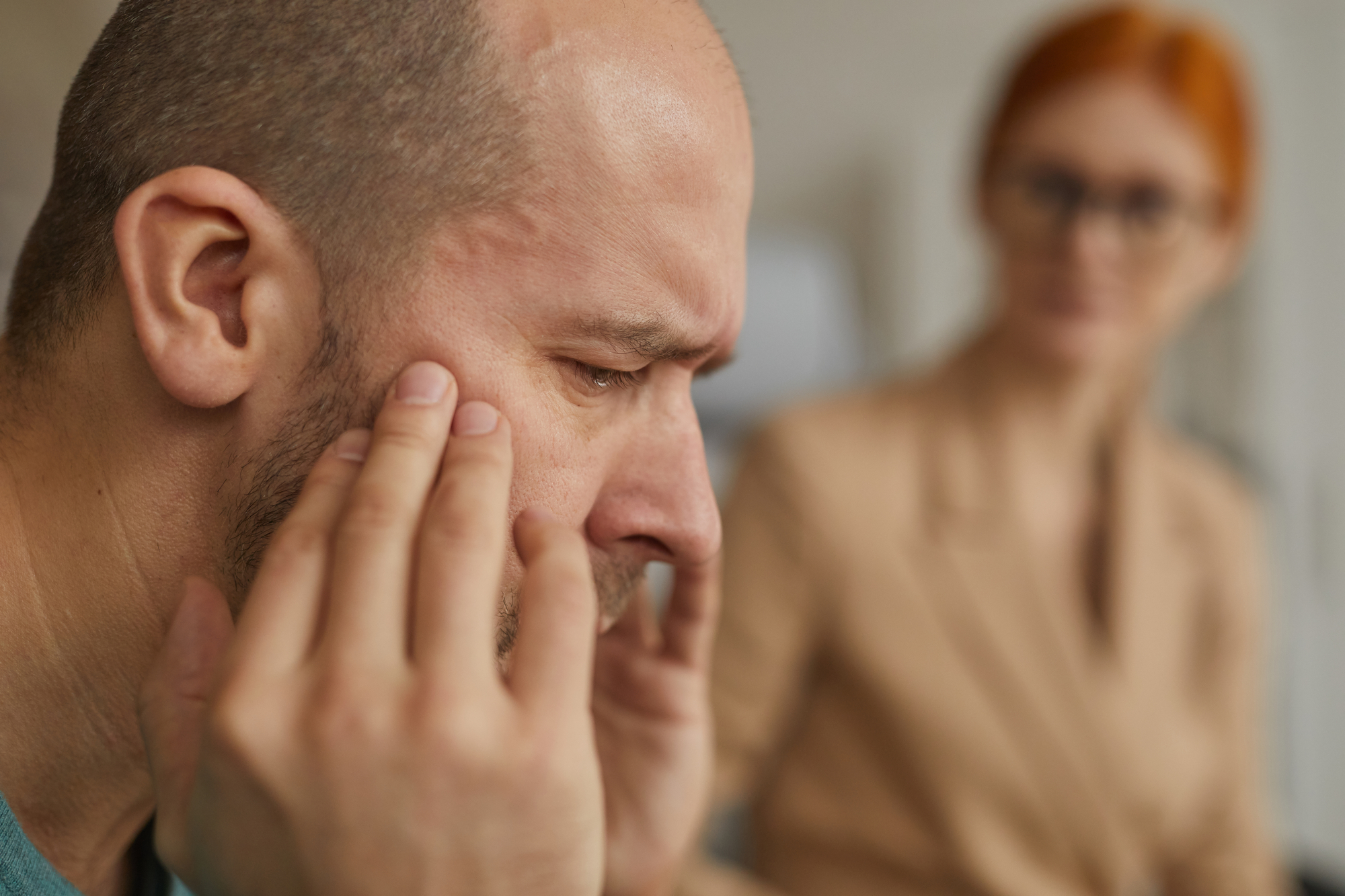 A man with a bald head and facial hair sits with his eyes closed and fingers on his temples, looking stressed or in pain. In the blurred background, a woman with red hair and glasses observes him attentively. Their expressions suggest concern and empathy.