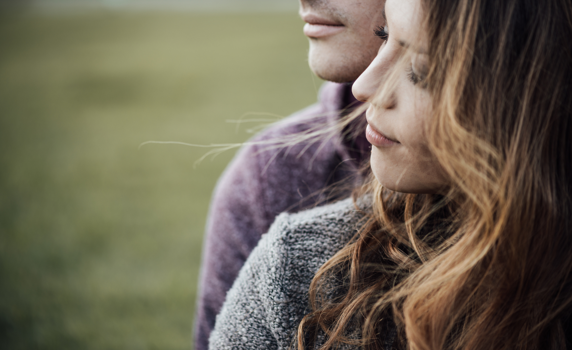 A close-up of a couple standing outdoors, with the woman in the foreground. She has long, wavy hair and wears a gray sweater. The man behind her wears a purple sweater. Both have serious expressions, and the background is a blurred green field.