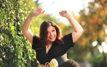 A woman with medium-length, dark hair is outdoors, smiling and posing with her hands raised above her head. She is wearing a black dress with a wide, gold belt. The background features sunlit, green foliage on the left and blurred trees with autumn colors on the right.
