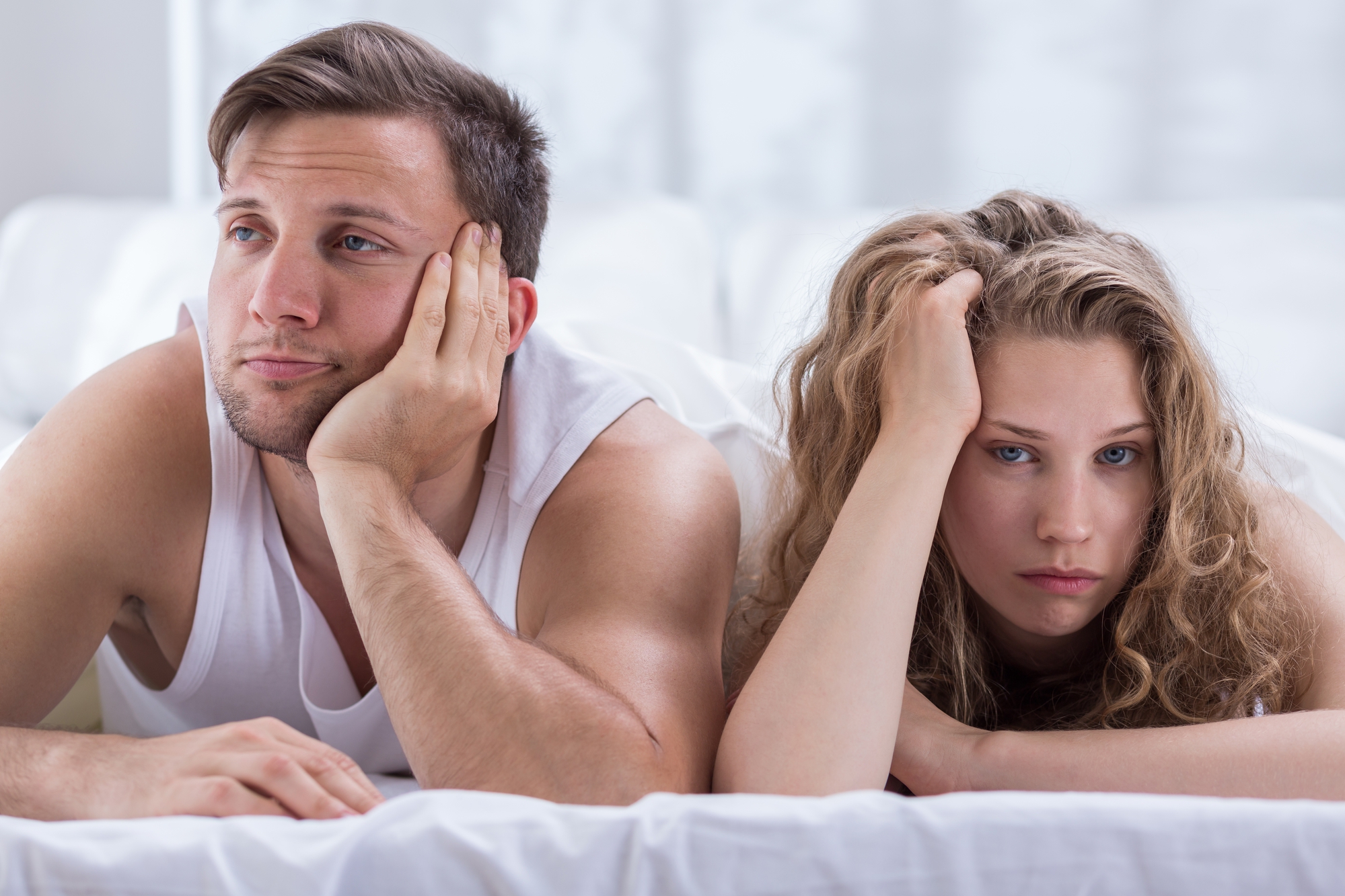 A man and a woman lie on a bed, resting their heads on their hands and looking bored or tired. Both have neutral or slightly unhappy expressions. The man is wearing a sleeveless shirt and the woman has curly hair. They appear to be in a bedroom setting.