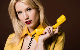 A woman with blonde hair and red lipstick holds a yellow retro-style corded phone to her ear while looking directly at the camera. She is wearing a yellow blazer, and the background is a dark brown color.
