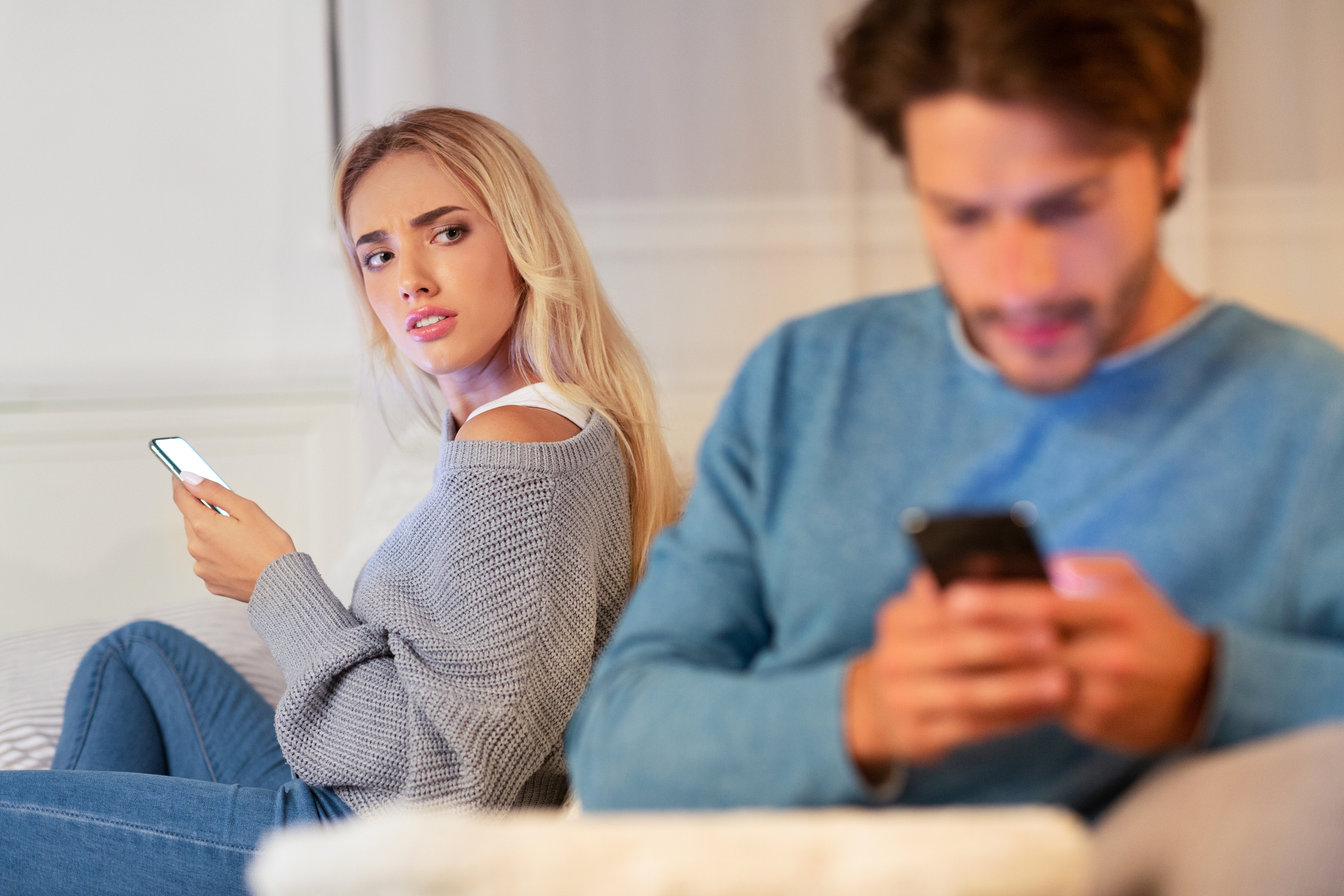 A woman with blonde hair, wearing a gray sweater, looks over at a man with brown hair, wearing a blue sweater. Both are sitting on a couch, each holding a smartphone. The woman appears puzzled or concerned as she glances toward the man, who is focused on his phone.