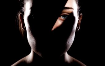 A woman's face is partially illuminated with a dramatic shadow obscuring most of her features, except for her left eye and part of her forehead. The background is black, creating a stark contrast with the light on her face.