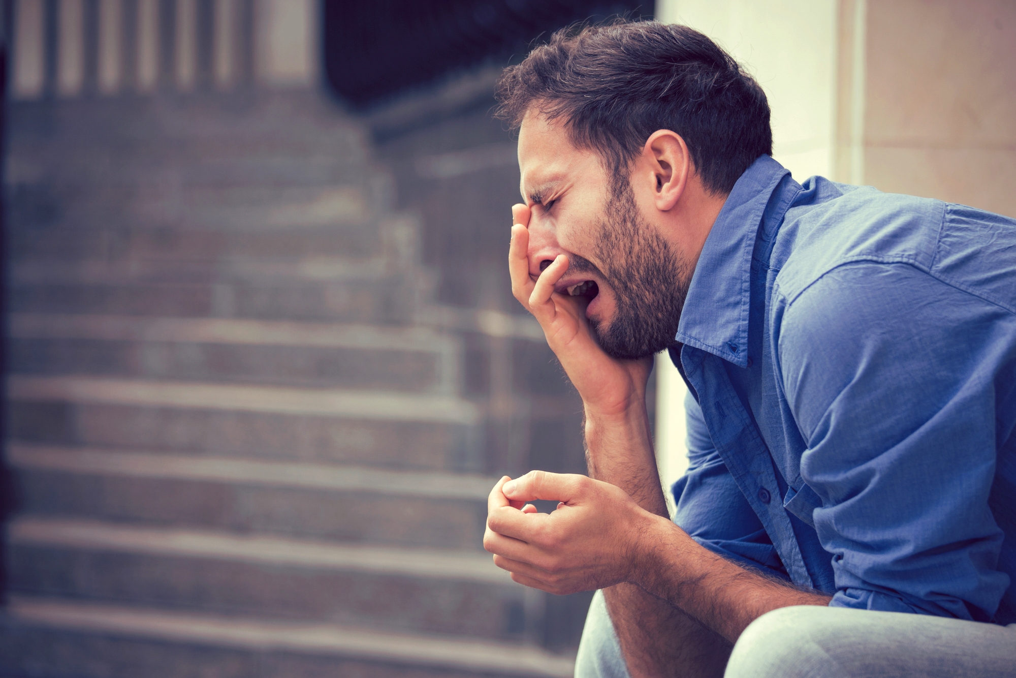 A man with a beard and wearing a blue shirt is sitting on steps, yawning or crying with one hand covering his face and the other hand extended out slightly. He appears to be tired or distressed.