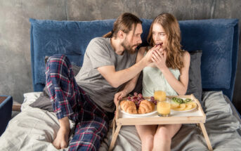 A man and woman are sitting on a bed with a blue headboard. The man, dressed in a grey t-shirt and plaid pants, is feeding the woman, who is wearing a light green camisole. A breakfast tray with croissants, fruits, and glasses of juice is placed on the bed.