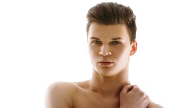 A young man with short, styled brown hair is against a plain white background. He is shirtless, with one hand resting on his shoulder. He has a serious expression on his face and is looking directly at the camera.