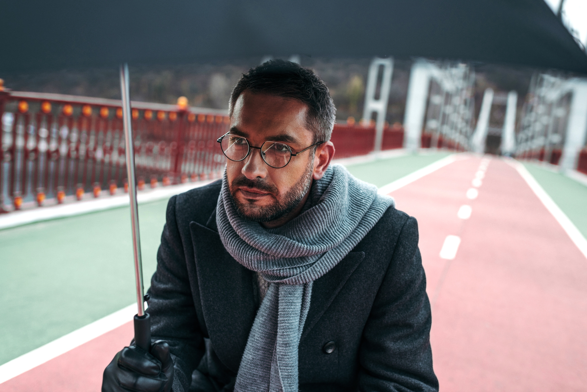 A man with glasses and a beard is sitting under a black umbrella on a bridge. He is wearing a dark coat and a gray scarf. The bridge has red railings and a green and red pathway with white lines. The background is slightly blurred, giving a sense of depth.